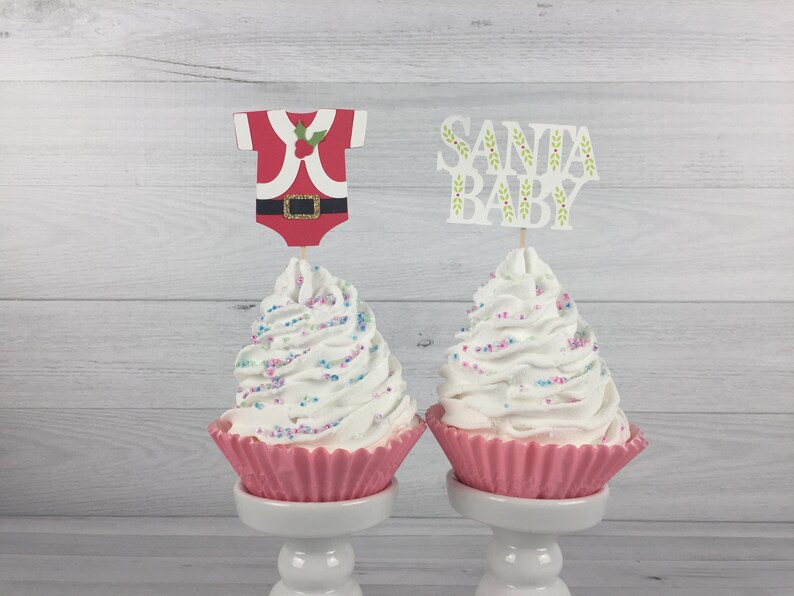 K&ouml;nnte beinhalten: Zwei rosa Cupcake-F&ouml;rmchen mit wei&szlig;em Frosting und bunten Streuseln. Ein Cupcake hat einen &bdquo;Santa Baby&ldquo;-Topper und der andere einen Santa-Baby-Strampler-Topper.