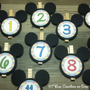 May include: Nine black Mickey Mouse head-shaped paper cutouts with colorful striped circles and numbers 1 through 8 and 11. Each cutout has a wooden clothespin attached.