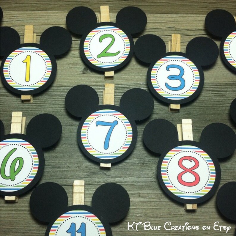 May include: Nine black Mickey Mouse head-shaped paper cutouts with colorful striped circles and numbers 1 through 8 and 11. Each cutout has a wooden clothespin attached.