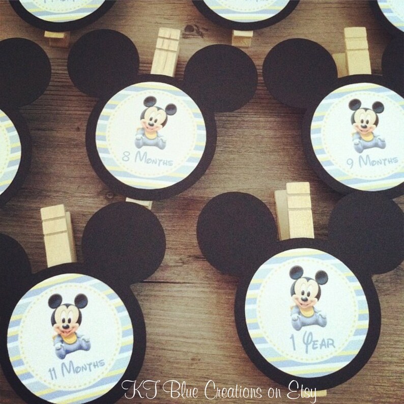 May include: Set of 5 black and white Mickey Mouse themed baby milestone photo props. Each prop features a black and white Mickey Mouse head with a yellow and blue striped circle in the center. The circles have a picture of Mickey Mouse in the center and text that says "8 Months", "9 Months", "11 Months", and "1 Year".