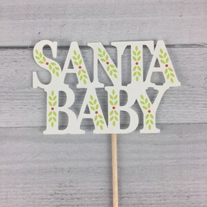 K&ouml;nnte beinhalten: Wei&szlig;er Tortendekor mit den Worten "Santa Baby" in einer festlichen Schriftart. Die Buchstaben sind mit gr&uuml;nen Bl&auml;ttern und roten Beeren verziert.