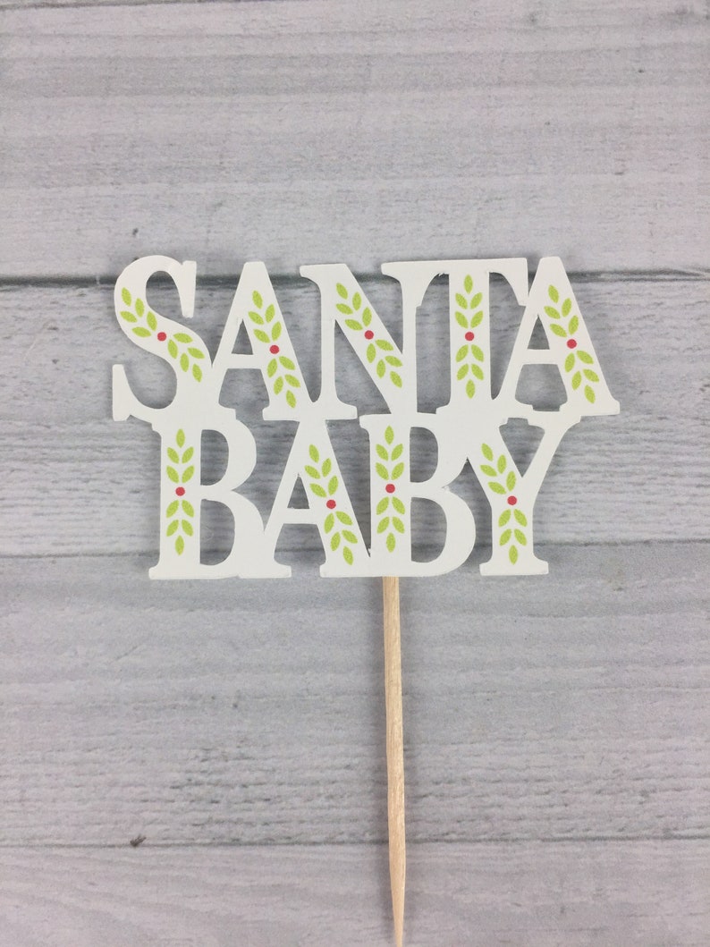 K&ouml;nnte beinhalten: Wei&szlig;er Tortendekor mit den Worten "Santa Baby" in einer festlichen Schriftart. Die Buchstaben sind mit gr&uuml;nen Bl&auml;ttern und roten Beeren verziert.