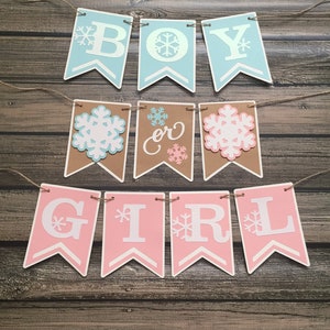 May include: A banner with the words "BOY or GIRL" in white capital letters on blue, brown, and pink pennants. Each pennant has a snowflake design. The banner is strung on twine and is set against a wooden background.