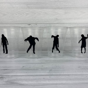 May include: Four clear plastic cups with black zombie silhouettes printed on them. The zombies are in various poses, including walking, running, and reaching out.