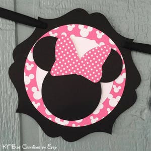 Op de afbeelding: Zwarte en roze Minnie Mouse-personagebanner met witte stippen. De banner heeft de vorm van een cirkel met een gegolfde rand.