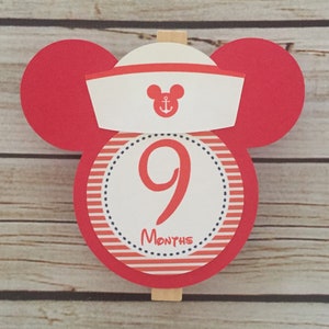May include: Red and white Mickey Mouse themed monthly milestone marker with a white sailor hat, an anchor, and the number 9. The text "MONTHS" is printed below the number.