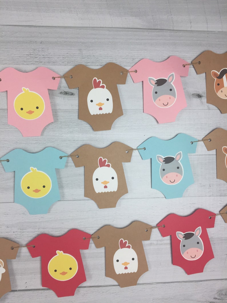 May include: A baby shower banner with a farm animal theme. The banner is made of brown, pink, blue, and red cardstock and features cutouts of baby onesies with animal faces. The animals include a chick, a chicken, and a donkey.