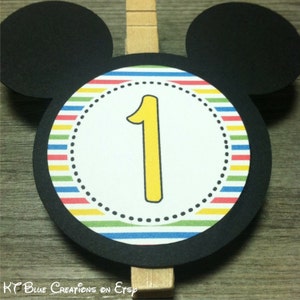 May include: Black Mickey Mouse head cutout with a colorful striped circle in the center. The circle has a white inner circle with a yellow number one in the center. The cutout is attached to a wooden clothespin.