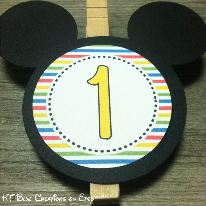May include: Black Mickey Mouse head cutout with a colorful striped circle in the center. The circle has a white inner circle with a yellow number one in the center. The cutout is attached to a wooden clothespin.