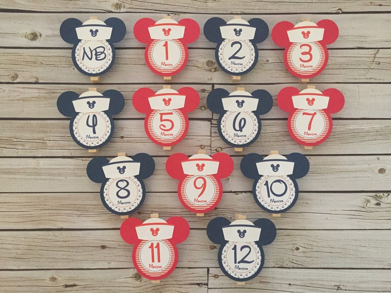 May include: Twelve red and blue Mickey Mouse-themed  paper clips with numbers 1 through 12 and "NB" printed on them. The clips are on a wooden background.