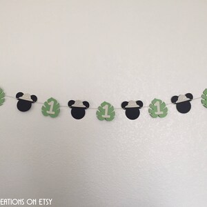 Puede incluir: Una pancarta de papel verde y negro con orejas de Mickey Mouse y el n&uacute;mero uno en cada hoja. La pancarta est&aacute; enhebrada con una cuerda blanca.