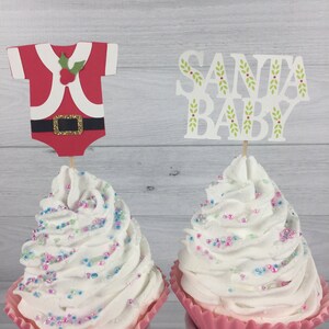 K&ouml;nnte beinhalten: Zwei Cupcakes mit wei&szlig;em Frosting und bunten Streuseln, die mit festlichen Weihnachts-Cupcake-Toppern verziert sind. Ein Topper ist ein rot-wei&szlig;es Weihnachtsmannkost&uuml;m und der andere sagt "Santa Baby".