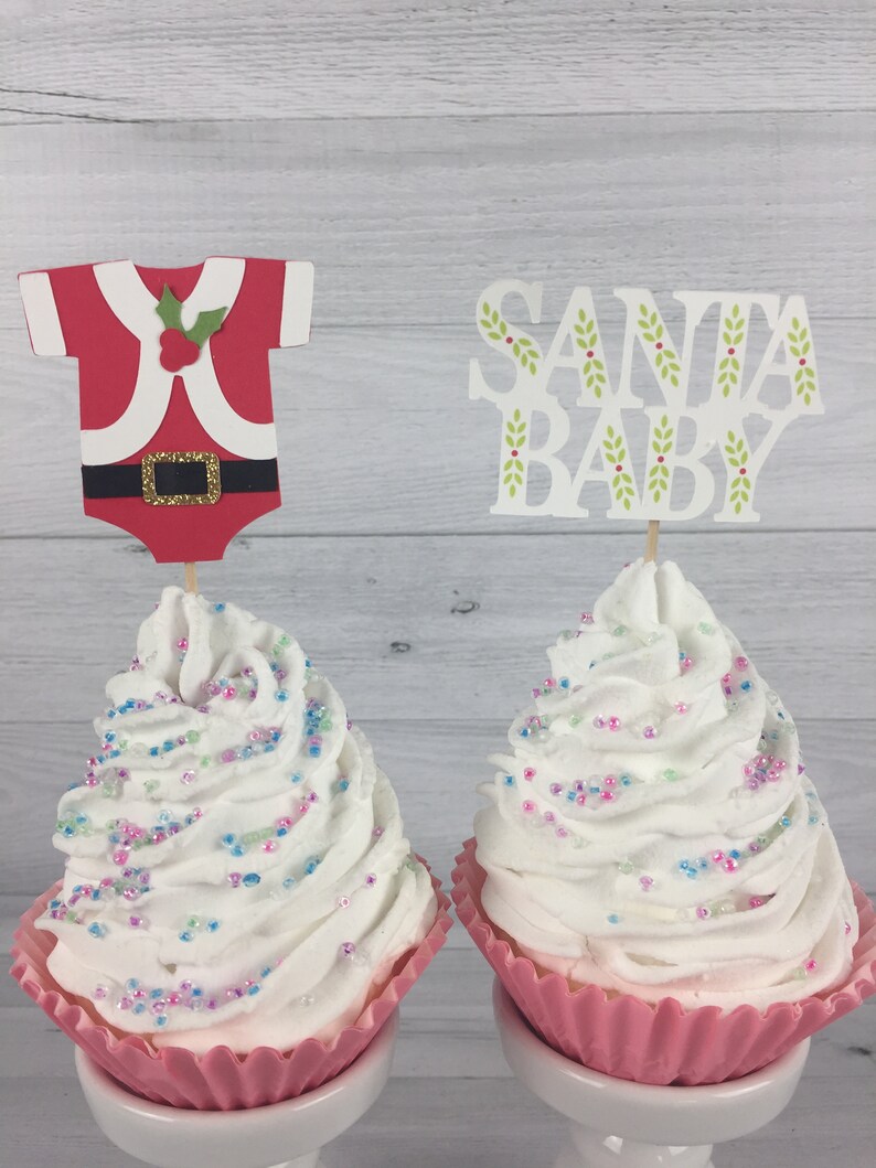 K&ouml;nnte beinhalten: Zwei Cupcakes mit wei&szlig;em Frosting und bunten Streuseln, die mit festlichen Weihnachts-Cupcake-Toppern verziert sind. Ein Topper ist ein rot-wei&szlig;es Weihnachtsmannkost&uuml;m und der andere sagt "Santa Baby".