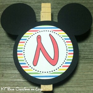 May include: Black and white Mickey Mouse-shaped card with a colorful striped circle in the center. The circle has a white background with a red letter "N" in the center. A wooden clothespin is attached to the top of the card.