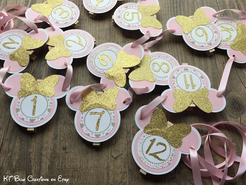 Op de afbeelding: Roze en gouden Minnie Mouse feestdecoraties met de nummers 1 tot en met 12. Elke decoratie heeft een roze Minnie Mouse hoofd met een gouden glitter strik en een witte cirkel met een gouden nummer.