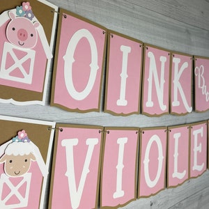 May include: Pink and brown banner with the text "Oink Moo Violet is 2" in white letters. The banner has a pig and a lamb with flowers on their heads in the corners.