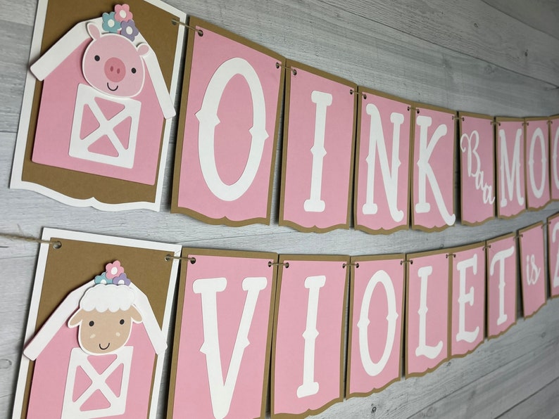 May include: Pink and brown banner with the text "Oink Moo Violet is 2" in white letters. The banner has a pig and a lamb with flowers on their heads in the corners.