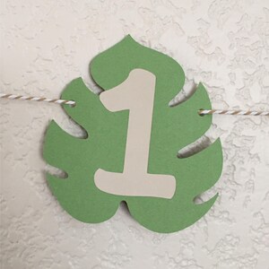 Puede incluir: Hoja verde de monstera con un n&uacute;mero uno blanco en ella. La hoja est&aacute; colgada de una cuerda.