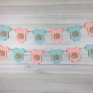 May include: A gender neutral baby shower banner with alternating pink and blue onesies decorated with gold turkey silhouettes.