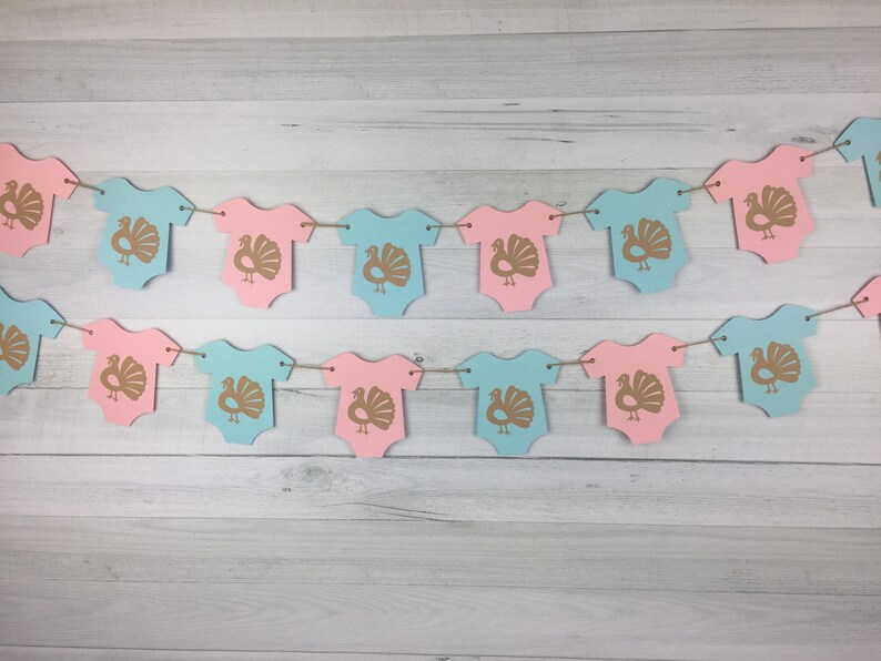 May include: A gender neutral baby shower banner with alternating pink and blue onesies decorated with gold turkey silhouettes.