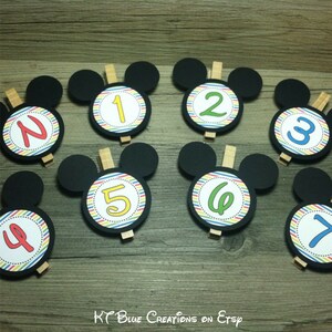 May include: Seven black Mickey Mouse head-shaped cardstock cutouts with colorful striped circles and numbers 1 through 7. The circles are attached to wooden clothespins.