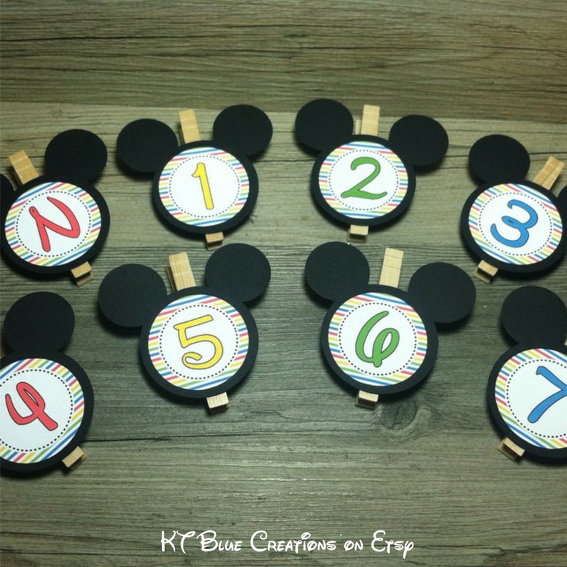 May include: Seven black Mickey Mouse head-shaped cardstock cutouts with colorful striped circles and numbers 1 through 7. The circles are attached to wooden clothespins.