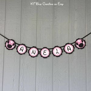 Op de afbeelding: Een zwart en roze banner met de naam "ANELA" gespeld in witte letters. Elke letter staat op een aparte cirkel met een roze stippenpatroon. De cirkels zijn verbonden door een zwart touwtje. Aan de uiteinden van de banner zitten twee zwarte en roze Minnie Mouse-koppen.