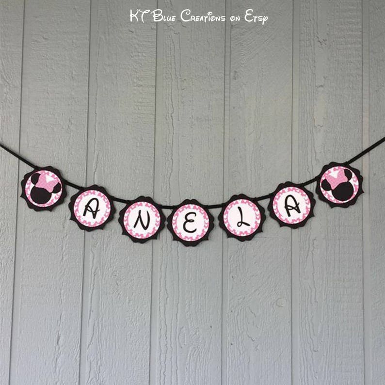 Op de afbeelding: Een zwart en roze banner met de naam "ANELA" gespeld in witte letters. Elke letter staat op een aparte cirkel met een roze stippenpatroon. De cirkels zijn verbonden door een zwart touwtje. Aan de uiteinden van de banner zitten twee zwarte en roze Minnie Mouse-koppen.