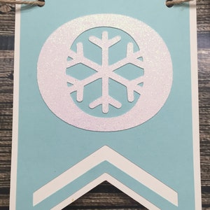 May include: A light blue banner with a white snowflake design. The snowflake is inside a white circle with glitter accents. The banner has a white border and a decorative bottom. A twine string is attached at the top.