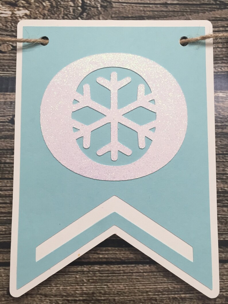 May include: A light blue banner with a white snowflake design. The snowflake is inside a white circle with glitter accents. The banner has a white border and a decorative bottom. A twine string is attached at the top.