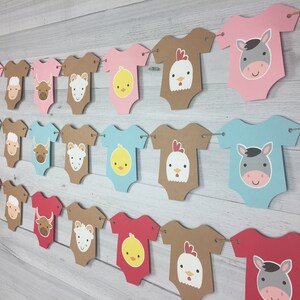 May include: A farm animal themed baby shower banner with brown, pink, blue, and red baby onesies with different animal faces. The banner is strung together with twine.