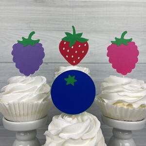 Peut inclure: Trois cupcakes avec du gla&ccedil;age blanc surmont&eacute;s de d&eacute;coupes en papier d'un bleuet, d'une fraise et d'une framboise. Les cupcakes sont sur des supports &agrave; g&acirc;teaux blancs.
