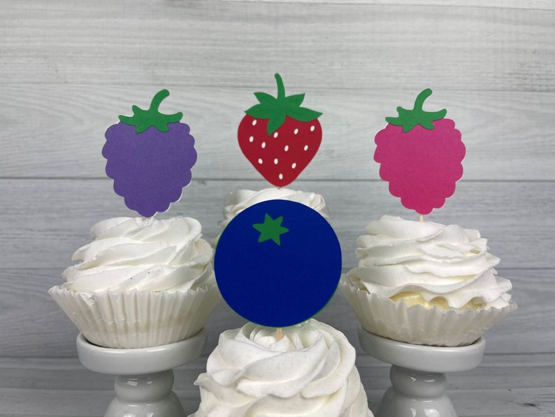 Peut inclure: Trois cupcakes avec du gla&ccedil;age blanc surmont&eacute;s de d&eacute;coupes en papier d'un bleuet, d'une fraise et d'une framboise. Les cupcakes sont sur des supports &agrave; g&acirc;teaux blancs.