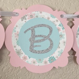 May include: Pink and blue snowflake patterned banner with a silver glitter letter B.
