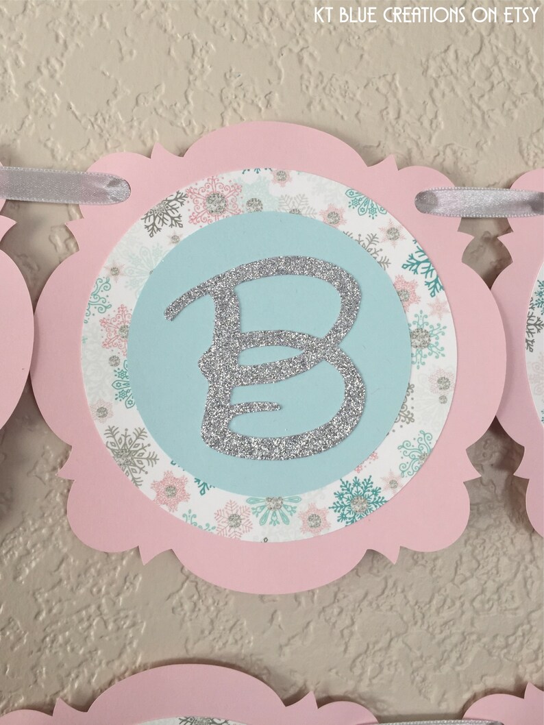 May include: Pink and blue snowflake patterned banner with a silver glitter letter B.