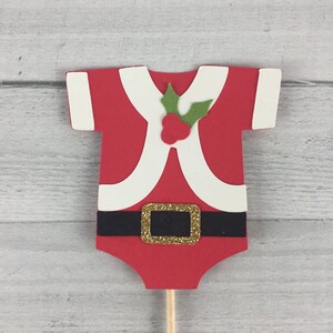 K&ouml;nnte beinhalten: Roter und wei&szlig;er Weihnachtsmann-Baby-Strampler-Kuchentopper mit einem gr&uuml;nen Stechpalmenblatt und roten Beeren. Der Strampler hat einen schwarzen G&uuml;rtel mit einer goldenen Schnalle.