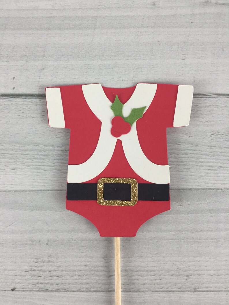 K&ouml;nnte beinhalten: Roter und wei&szlig;er Weihnachtsmann-Baby-Strampler-Kuchentopper mit einem gr&uuml;nen Stechpalmenblatt und roten Beeren. Der Strampler hat einen schwarzen G&uuml;rtel mit einer goldenen Schnalle.