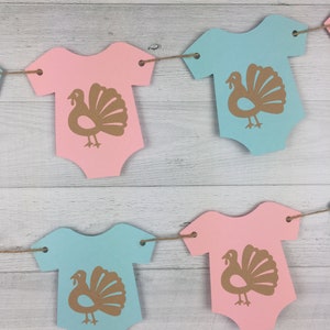 May include: A gender neutral baby shower banner with alternating pink and blue baby onesies with a gold turkey silhouette on each onesie.