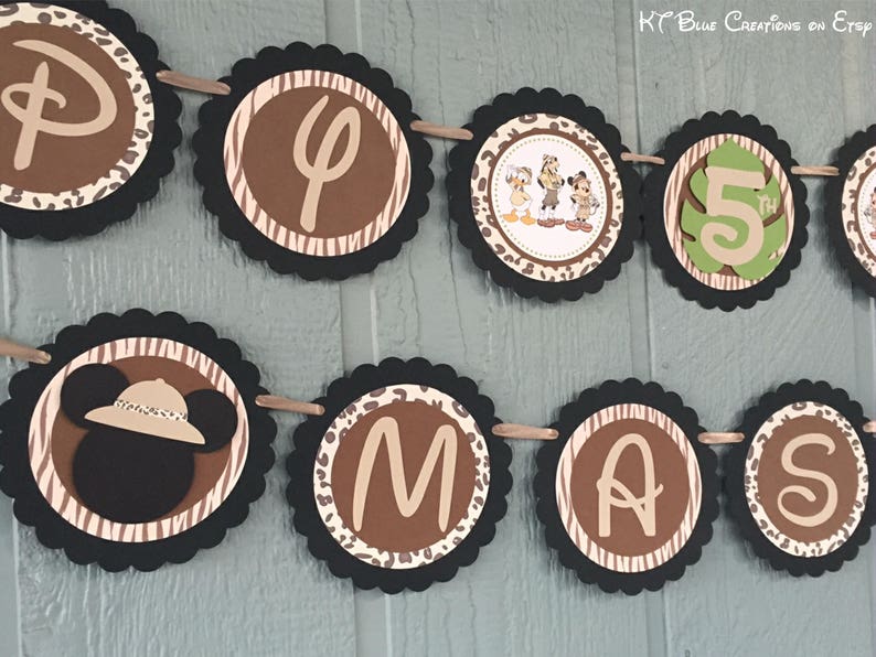 May include: A black and brown jungle-themed banner with the letters "D-I-A-M-A-S" and a number "5" in a green leaf. The banner also features a cartoon image of Mickey Mouse wearing a safari hat and Donald Duck and Goofy.