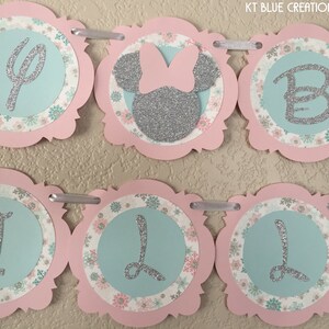 May include: A pink and blue banner with silver glitter letters spelling "It's a Girl". The banner has a floral pattern and a silver glitter Minnie Mouse head with a pink bow.