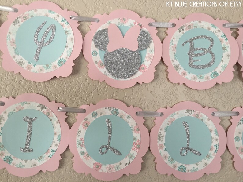 May include: A pink and blue banner with silver glitter letters spelling "It's a Girl". The banner has a floral pattern and a silver glitter Minnie Mouse head with a pink bow.