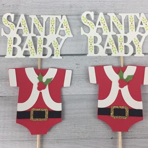 K&ouml;nnte beinhalten: Zwei &bdquo;Santa Baby&ldquo;-Kuchentopper. Jeder Topper zeigt einen roten und wei&szlig;en Weihnachtsmann-Anzug mit einem gr&uuml;nen Stechpalmenzweig und einer goldenen Schnalle. Oben auf jedem Topper befindet sich ein wei&szlig;es Schild mit der Aufschrift &bdquo;Santa Baby&ldquo; mit gr&uuml;nen Bl&auml;ttern und roten Beeren.