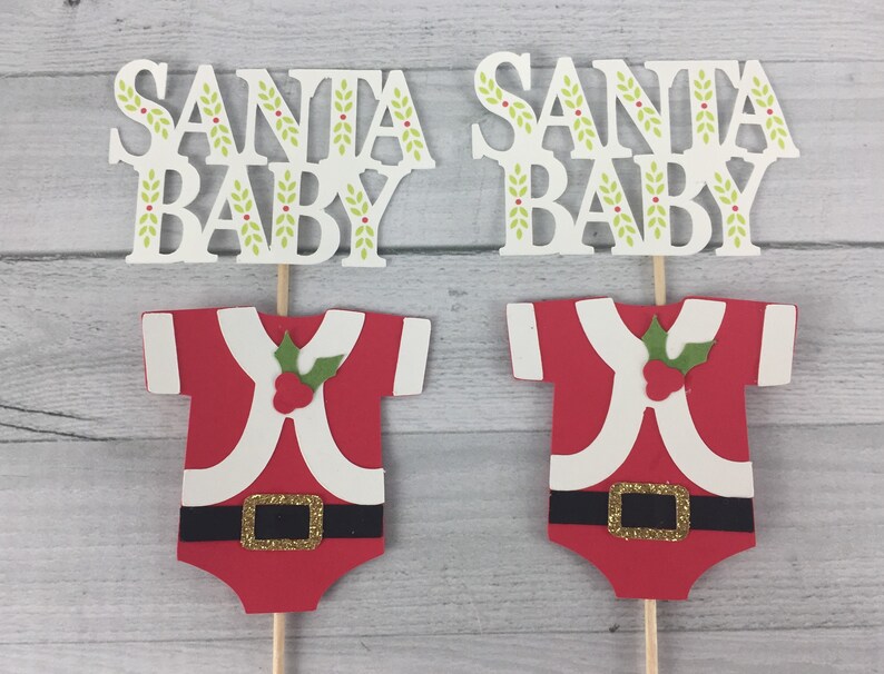 K&ouml;nnte beinhalten: Zwei &bdquo;Santa Baby&ldquo;-Kuchentopper. Jeder Topper zeigt einen roten und wei&szlig;en Weihnachtsmann-Anzug mit einem gr&uuml;nen Stechpalmenzweig und einer goldenen Schnalle. Oben auf jedem Topper befindet sich ein wei&szlig;es Schild mit der Aufschrift &bdquo;Santa Baby&ldquo; mit gr&uuml;nen Bl&auml;ttern und roten Beeren.