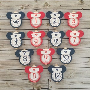 May include: Twelve red and blue Mickey Mouse-shaped cards with numbers 1 through 12 and the word "Months" printed on each card. The cards are attached to wooden clothespins.