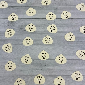 May include: A collection of beige paper cutouts of various facial expressions, resembling steamed buns, arranged on a gray wood background.
