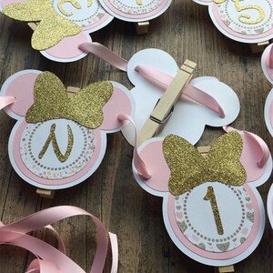 Op de afbeelding: Roze en gouden Minnie Mouse-banner met de nummers 1 tot en met 5. Elk nummer staat op een cirkel met een roze en gouden glitter strik en een roze en gouden stippenpatroon. De banner is aan elkaar geregen met roze lint.