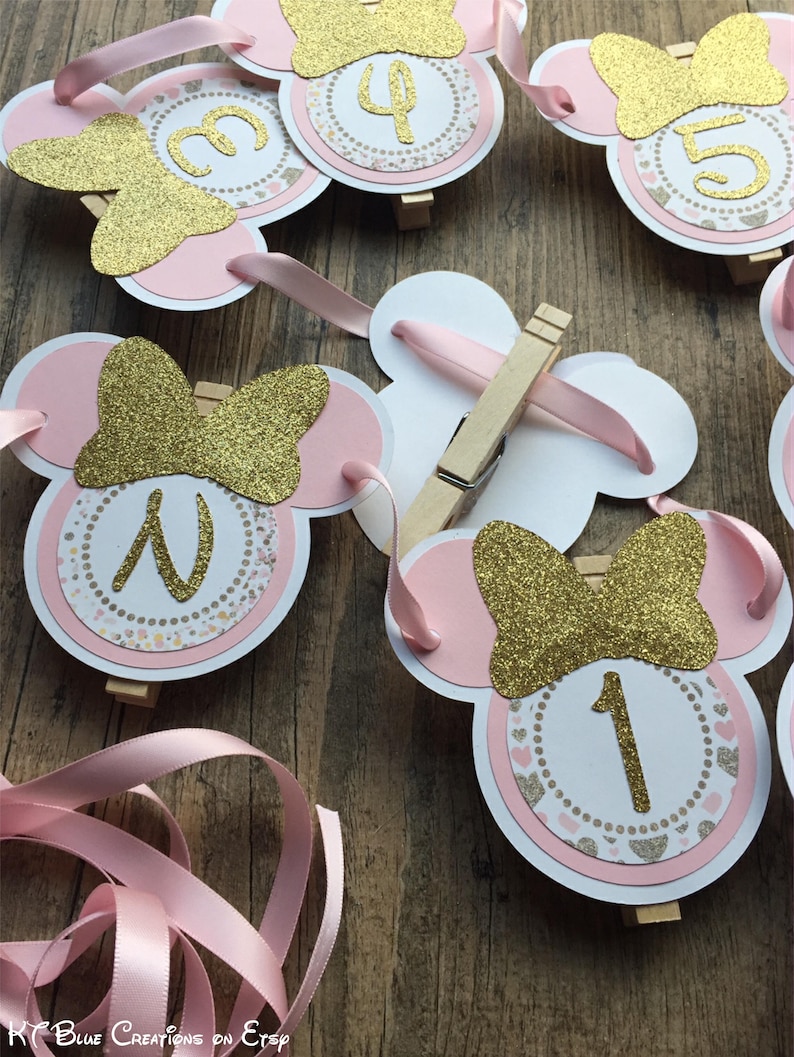 Op de afbeelding: Roze en gouden Minnie Mouse-banner met de nummers 1 tot en met 5. Elk nummer staat op een cirkel met een roze en gouden glitter strik en een roze en gouden stippenpatroon. De banner is aan elkaar geregen met roze lint.
