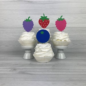 Peut inclure: Quatre cupcakes avec du gla&ccedil;age blanc et des garnitures de fruits diff&eacute;rentes. Les garnitures sont un raisin violet, une fraise rouge, une framboise rose et une myrtille bleue.