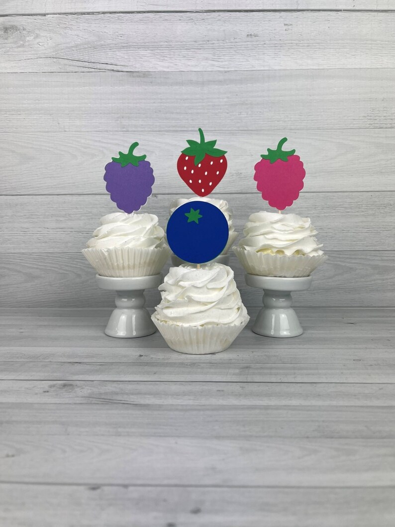 Peut inclure: Quatre cupcakes avec du gla&ccedil;age blanc et des garnitures de fruits diff&eacute;rentes. Les garnitures sont un raisin violet, une fraise rouge, une framboise rose et une myrtille bleue.
