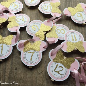 Op de afbeelding: Roze en gouden Minnie Mouse feestdecoraties met de nummers 1 tot en met 12. Elke decoratie heeft een roze Minnie Mouse hoofd met een gouden glitter strik en een witte cirkel met een gouden nummer.